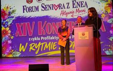 Kobieta w czarnej koszuli i czerwonych spodniach przemawia przy m&oacute;wnicy. Z tyłu stoi mężczyzna w garniturze i kobieta w spodniach i kolorowej kosuli. W tle wyświetlony r&oacute;żowy napis "XIV Konferencja z cyklu "Profilaktycznie w Połańcu. Forum serniora z  Eneą" 