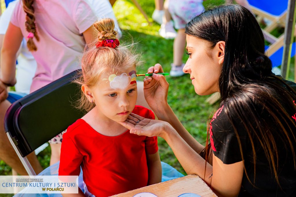 Na zdjęciu kobieta malująca tęczę na twarzy małej dziewczynki