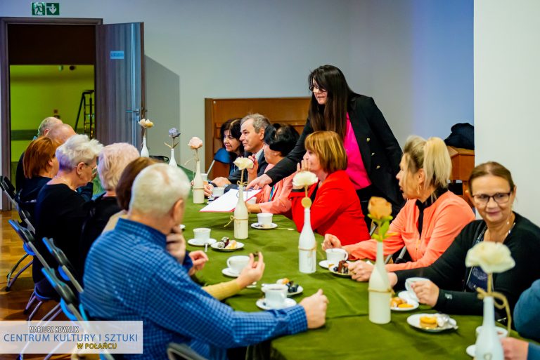 Na zdjęciu grupa siedzących przy stole starszych osób i stojąca przy nich kobieta