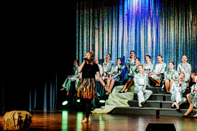 Widowisko taneczno-muzyczne "Mam tę moc" - śpiewająca piosenkę grupa chórzystów siedząca na schodach, z przodu śpiewająca dziewczyna