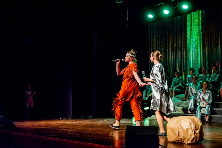 Widowisko taneczno-muzyczne "Mam tę moc" - śpiewająca piosenkę grupa chórzystów siedząca na schodach, z przodu dwie Indianki i dziewczynka w srebrnej sukience