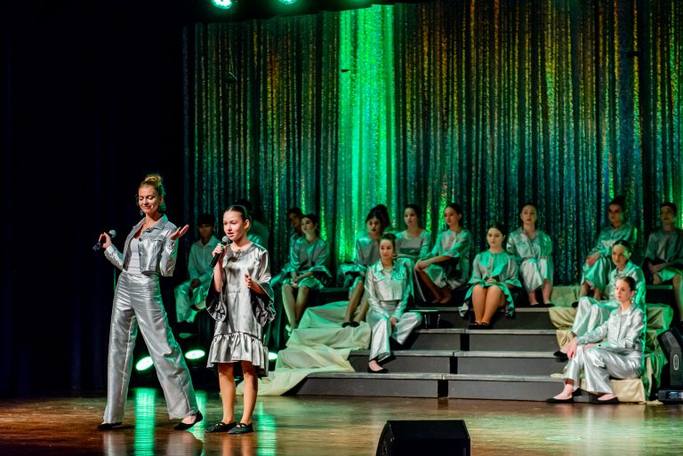 Widowisko taneczno-muzyczne "Mam tę moc" - śpiewająca piosenkę grupa chórzystów siedząca na schodach, z przodu tańczy dziewczynka w srebrnej sukience i dziewczyna w srebrnym stroju