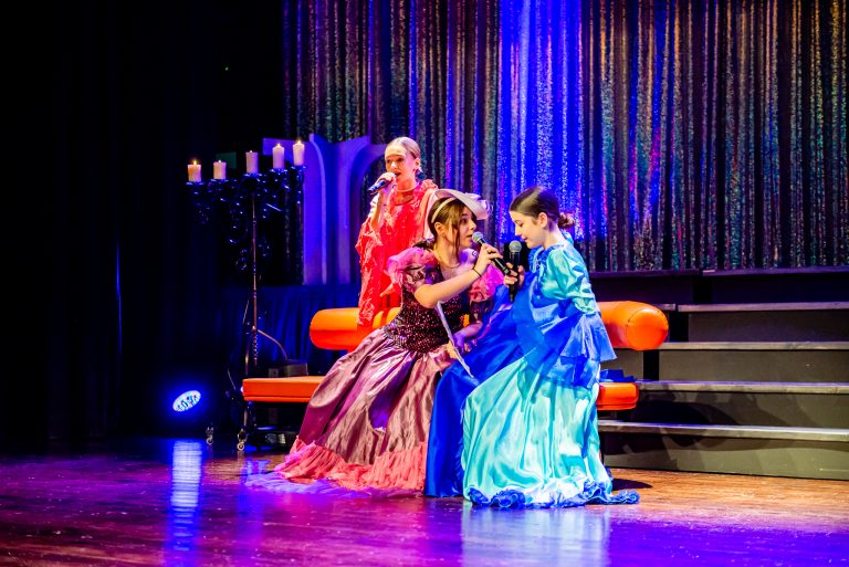 Widowisko taneczno-muzyczne "Mam tę moc" - trzy śpiewające dziewczyny ubrane w suknie, siedzą na pomarańczowej kanapie