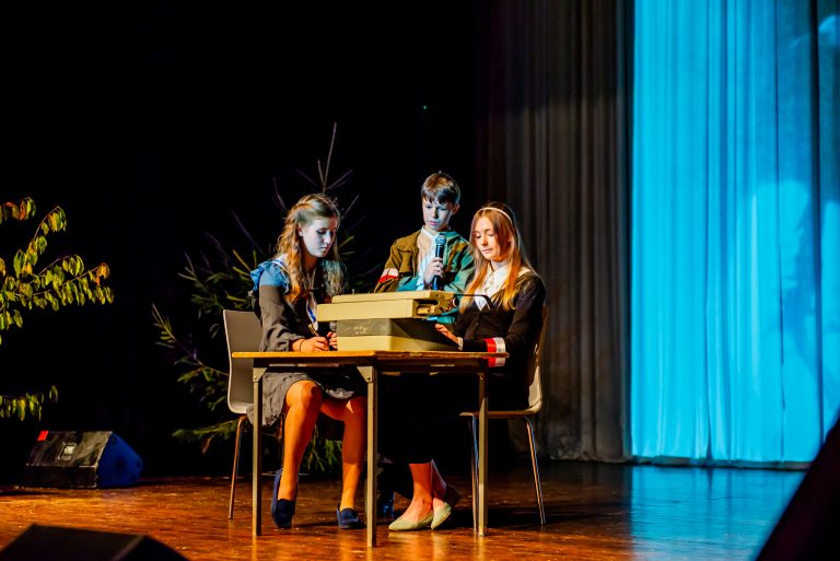 Na zdjęciu grupa młodych ludzi znajdująca się na scenie , odgrywająca scenę ze spektaklu - dwie dziewczyny siedzą przy stoliku, chłopiec stoi przy nich