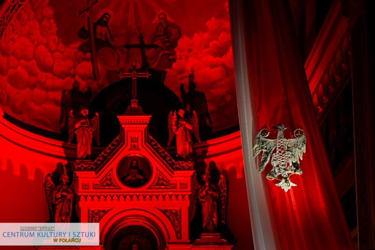Zdjęcie przedstawia flagę Polski, na której widnieje figurka orła, w tle ołtarz