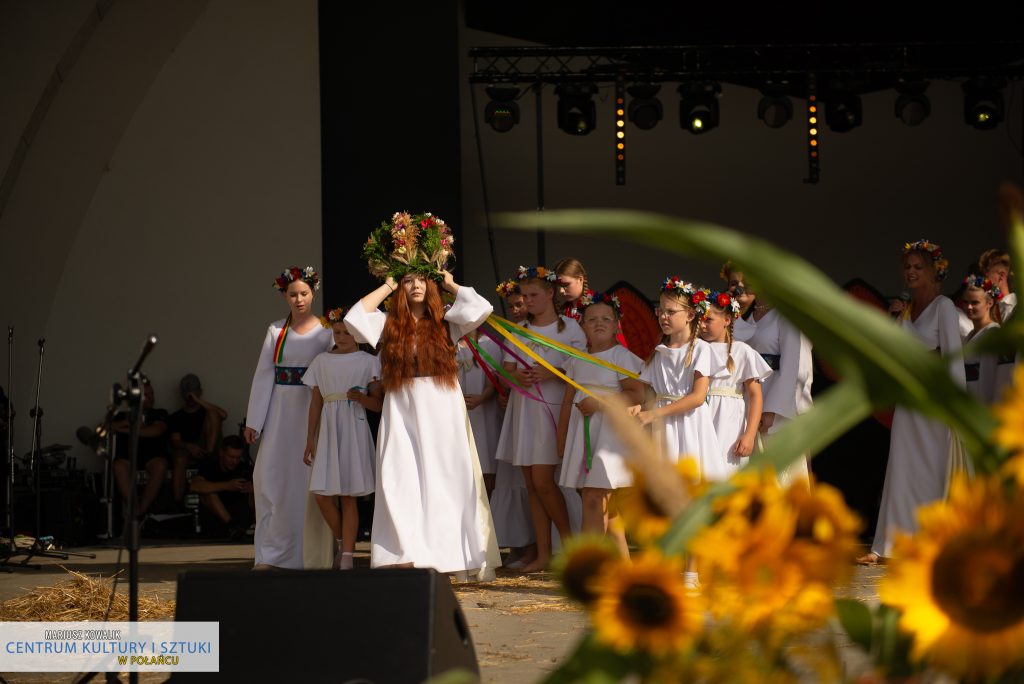 Scena z "Misterium Żniwnego" - Dziewczyna z długimi, rudymi włosami, niosąca na głowie koronę w formie wieńca dożynkowego. Wstążki doczepione do wieńca trzymają małe dziewczynki. Z tyłu idzie grupa dziewcząt