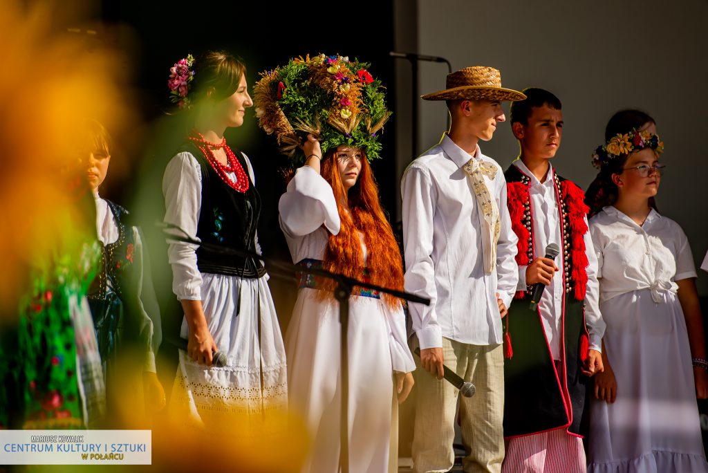 Scena z "Misterium Żniwnego" - Na środku zdjęcia jest młoda dziewczyna w długim, rudych włosach z koroną w formie wieńca dożynkowego. Obok niej po lewej stronie stoi gospodyni, po prawej gospodarz, chłopiec i dziewczyna. 