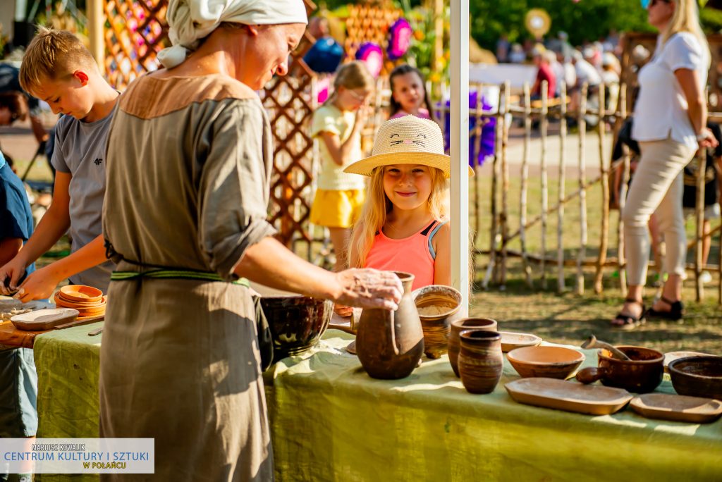 Na zdjęciu stoisko z naturalną ceramiką, obsługujący przez panią ubraną w lnianą sukienkę. Przy stoisku stoi uśmiechnięta dziewczynka w różowej sukience i słomkowym kapeluszu oraz chłopiec w szarej koszulce.