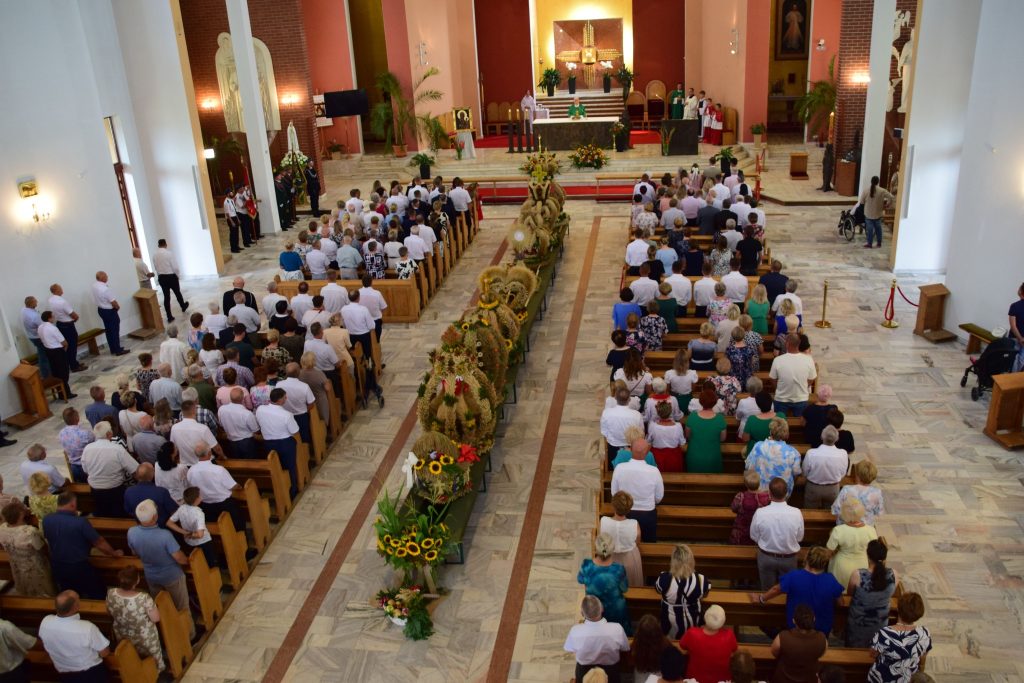 Widok z góry na główną nawę kościoła. Po środku ustawione są w rzędzie wieńce dożynkowe. Ławki wypełnione są elegancko ubranymi ludźmi. 