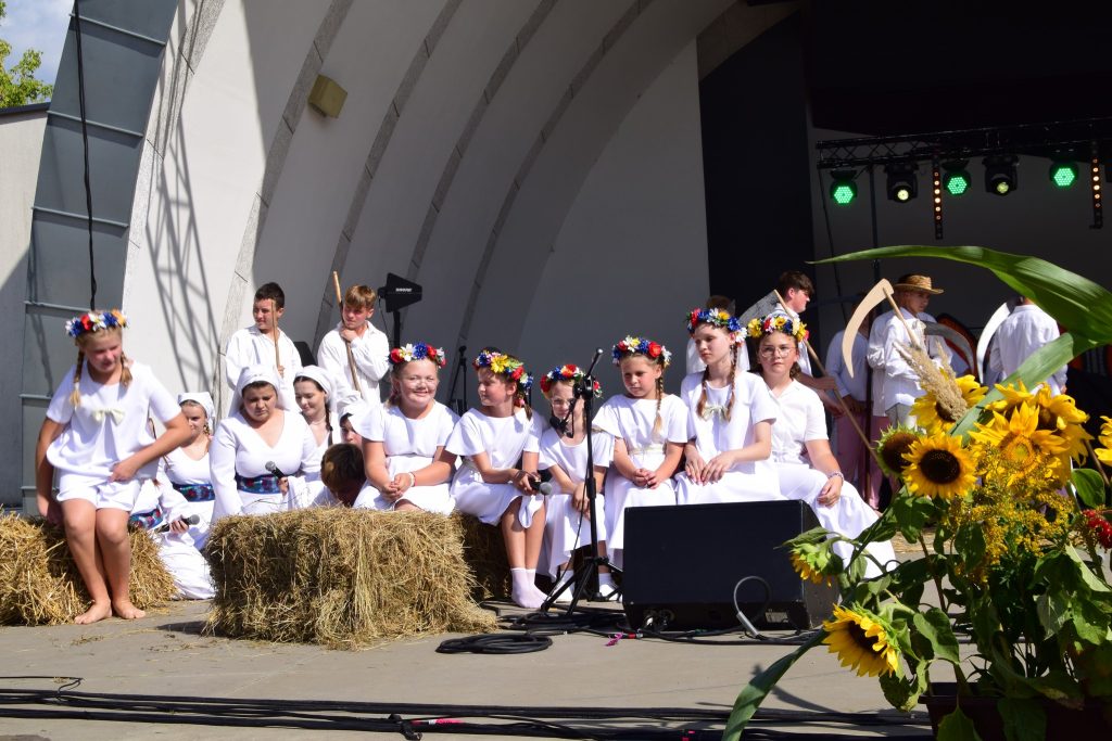 Scena z "Misterium Żniwnego" - Grupa małych dziewczynek i młodych dziewcząt, siedząca na snopach. Z tyłu stoją chłopacy oparci o kosy, imitujący odpoczynek po pracy w polu