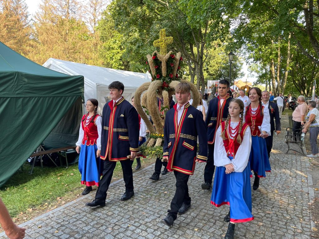 Czterech chłopców niosących wieniec dożynkowy oraz grupa reprezentacyjna idąca przy wieńcu - dziewczęta ubrane w ludowe stroje 