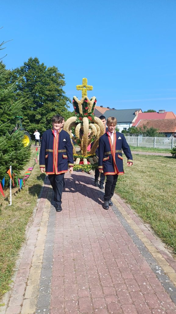 Czterech chłopców w granatowych marynarkach z czerwonym obszyciem niesie wieniec dożynkowy gminy Połaniec