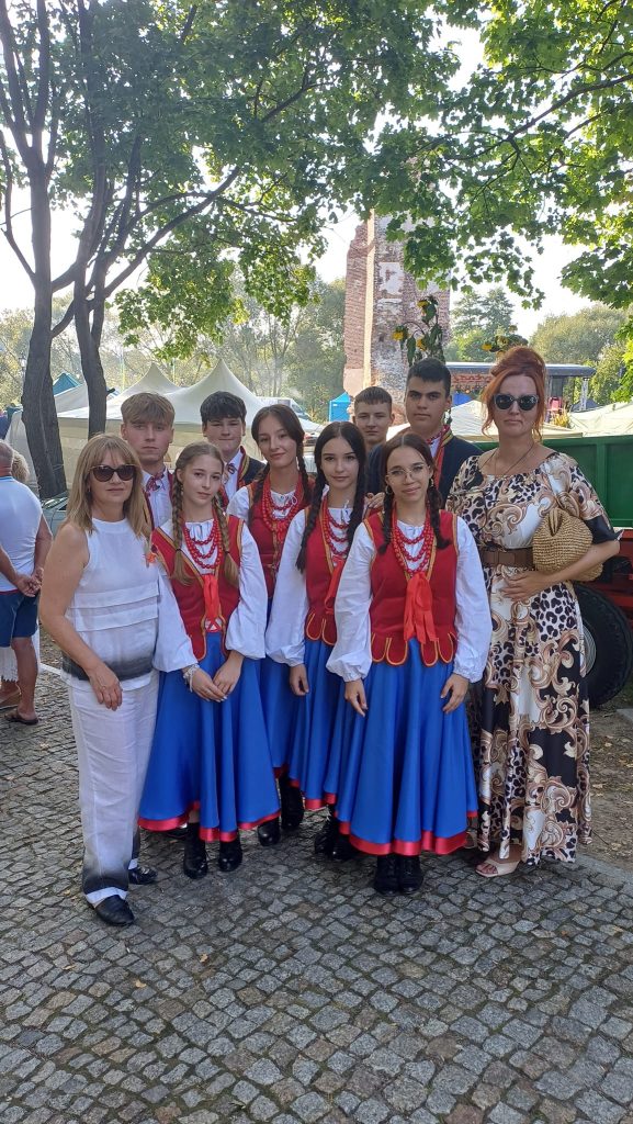 Na zdjęciu grupa reprezentacyjna CKiSz - dyrektorka CKiSz, instruktor zajęć teatralnych oraz grupa teatralna "Maska" - cztery dziewczęta ubrane w ludowy strój (niebieską spódnicę, białe koszule oraz czerwone wiązane gorsety i czerwone korale) oraz czterech chłopców ustawionych za dziewczętami